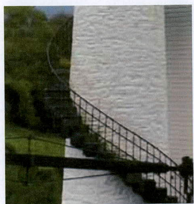 A close-up photograph showing a metal railing for an external staircase running alongside a white rendered wall, with greenery visible to the left.
