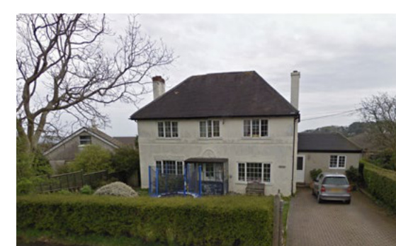A photograph showing a white two-story detached house with a dark roof, situated in a rural setting with hedges and a driveway.