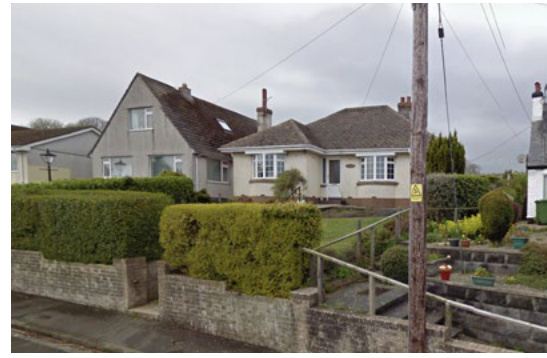 A street-level photograph showing a cream-colored bungalow and a larger grey house situated behind a stone wall and hedge in a rural setting.