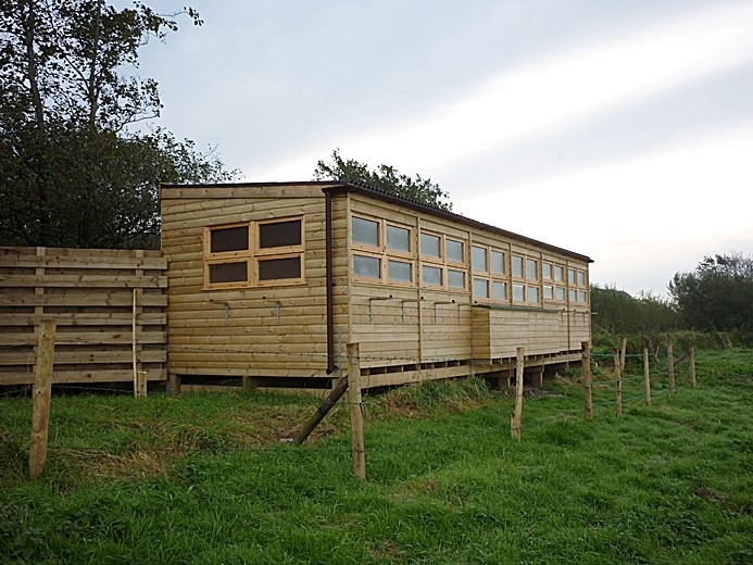 A photograph showing a long, timber-framed structure raised on wooden stilts in a grassy field. The building features a row of windows along the side, consistent with a birdwatching hide.