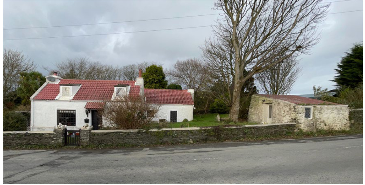 Street-level photograph of a white single-story dwelling with a red tiled roof and an adjacent stone outbuilding, separated from the road by a low stone wall.