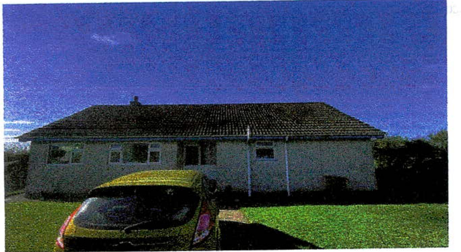 A grainy photograph showing a single-story detached bungalow with a tiled roof and light-colored walls. A yellow car is parked in the foreground on a grassy area.