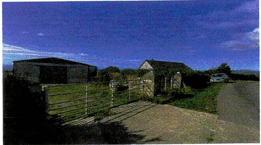 A grainy photograph showing a rural property with a large agricultural-style building on the left and a smaller house on the right, viewed through a metal gate.