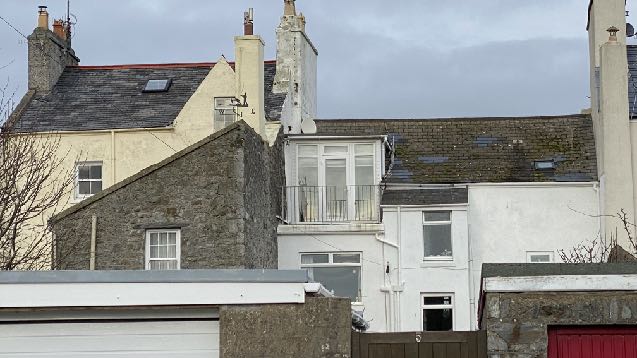 A street-level photograph showing the rooflines and upper facades of a row of residential buildings, including a stone house and a white building with a bay window.