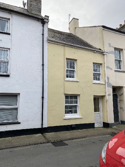 A street-level photograph showing a row of terraced buildings, featuring a white building on the left and a yellow two-story house in the center.