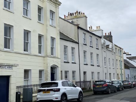A street-level photograph showing a row of multi-story terraced buildings with traditional facades and chimneys. Several cars are parked along the curb in front of the properties.