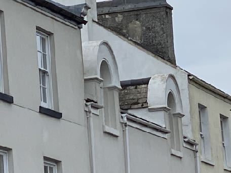 A close-up photograph of the upper facade and roofline of a white rendered terraced house, showing the chimney stack, guttering, and decorative arches.