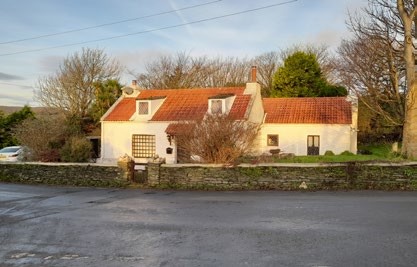A street-level photograph of a white, single-story detached house with a red tiled roof and dormer windows, situated behind a stone wall.
