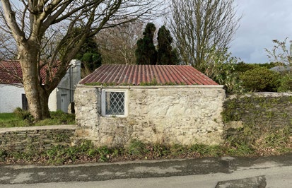 A photograph showing a small stone outbuilding with a corrugated roof and a single window, situated beside a road with a stone wall.
