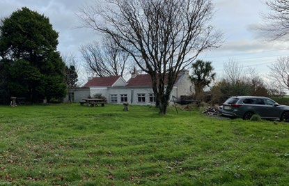 A photograph showing a white, single-story detached house with a red roof situated in a grassy garden with trees and a parked car.