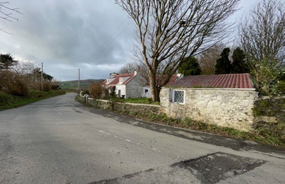 A street-level photograph showing a rural road with stone buildings, including a small detached garage and a larger white house, set against a cloudy sky.
