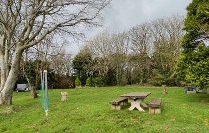 A photograph showing a grassy garden area with a wooden picnic table, benches, and a swing set surrounded by trees.