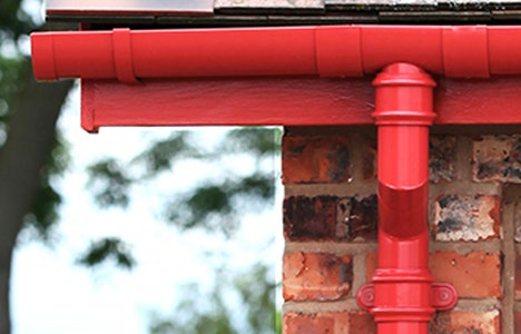 A close-up photograph showing a red plastic gutter and downpipe attached to a red brick wall, with blurred greenery in the background.