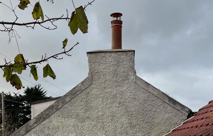 A photograph showing the white rendered gable end of a building with a chimney stack and terracotta pot, framed by tree branches against a cloudy sky.