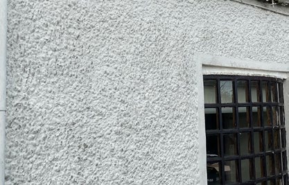 A close-up photograph of a white pebbledash exterior wall featuring a black-framed window with a grid pattern.