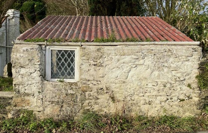A photograph of a small, single-story stone outbuilding with a corrugated roof and a diamond-paned window.