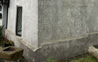 A close-up photograph of the exterior corner of a house featuring roughcast rendering, a diamond-paned window, and a stone trough.