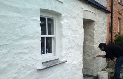 A close-up photograph showing a person painting or rendering the white-washed stone exterior wall of a building next to a white window.