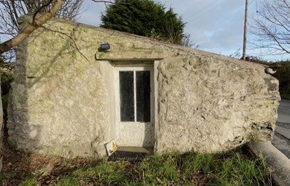 A photograph showing the exterior of a small, single-story stone building with a white door, surrounded by vegetation and trees.