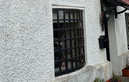 A close-up photograph of a white pebbledash exterior wall featuring a window with black security bars and a mounted black mailbox.