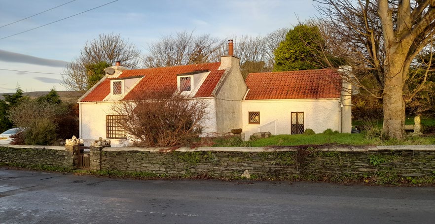 A photograph of a white, single-story detached dwelling with a red tiled roof and dormer windows. The property is situated behind a low stone wall with mature trees and shrubbery.