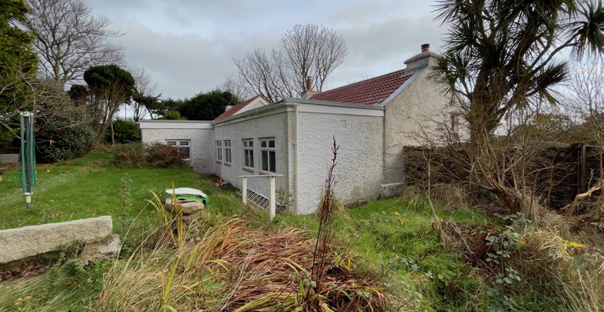 A photograph of a white rendered bungalow featuring a single-story extension with large windows, set in a grassy garden area.