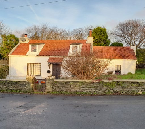 A photograph of a white, single-story detached house with a red tiled roof and dormer windows. The property is fronted by a low stone wall and situated in a rural setting.