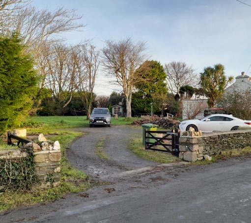 A photograph showing the rural site entrance with a gravel driveway, stone boundary walls, and a black gate, with vehicles parked on the property.