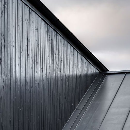 A close-up photograph showing the corner of a building featuring dark vertical timber cladding and a sloping roof against an overcast sky.