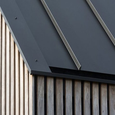 A close-up photograph of a building exterior corner featuring vertical timber cladding and a dark grey metal roof or fascia.