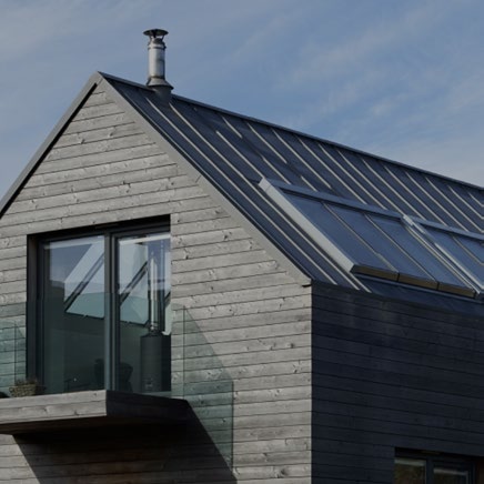 A close-up view of a modern building's upper facade and roof, featuring grey timber cladding, a metal roof with skylights or solar panels, and a large window.