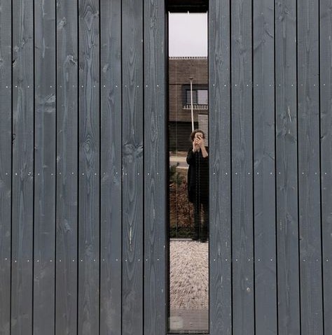 A close-up photograph of a dark grey vertical wooden slatted structure with a narrow vertical opening revealing a person and building behind.