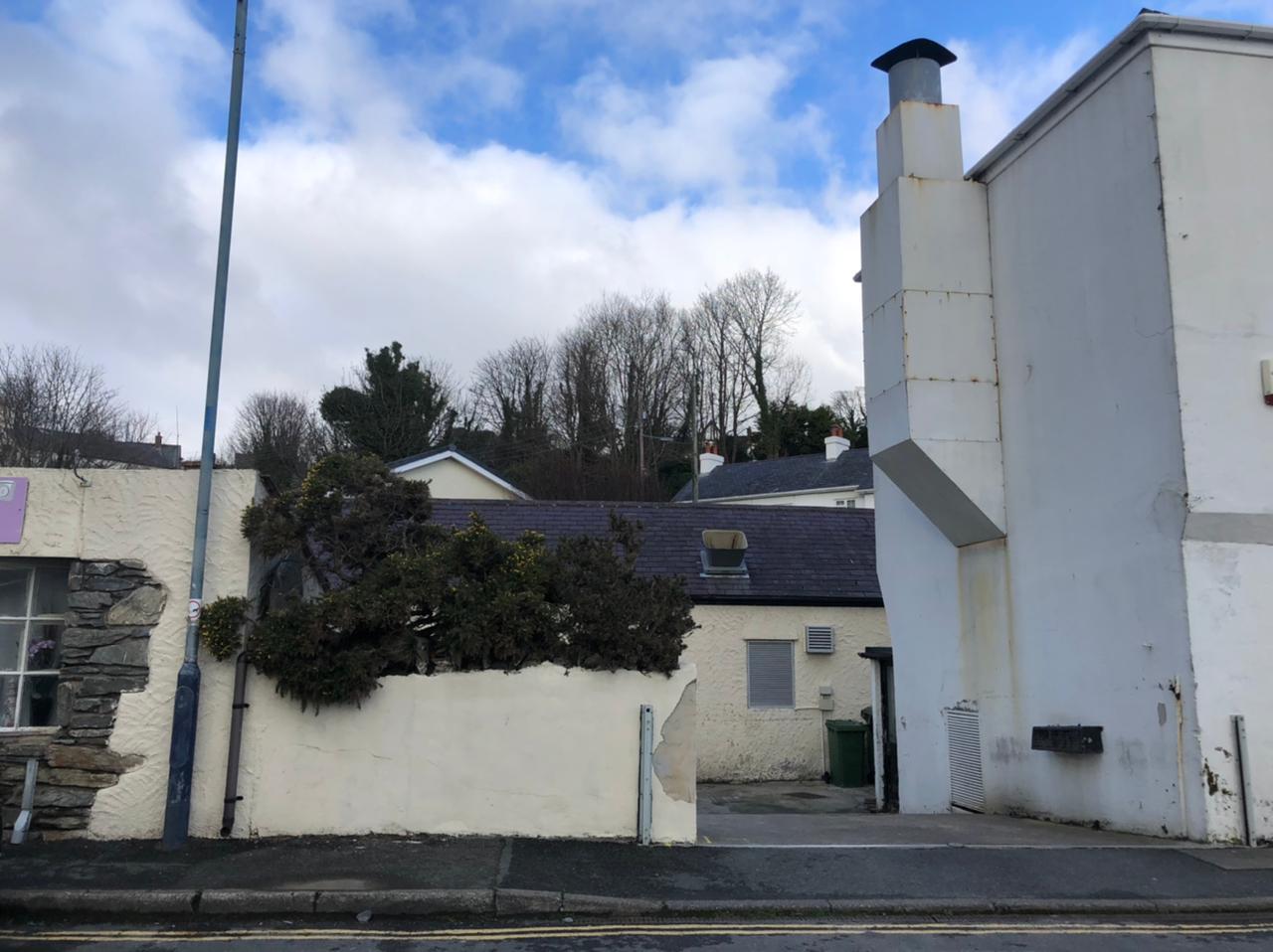 A street-level photograph showing a white rendered boundary wall with a gap, adjacent to a building with a slate roof and a tall white chimney structure.