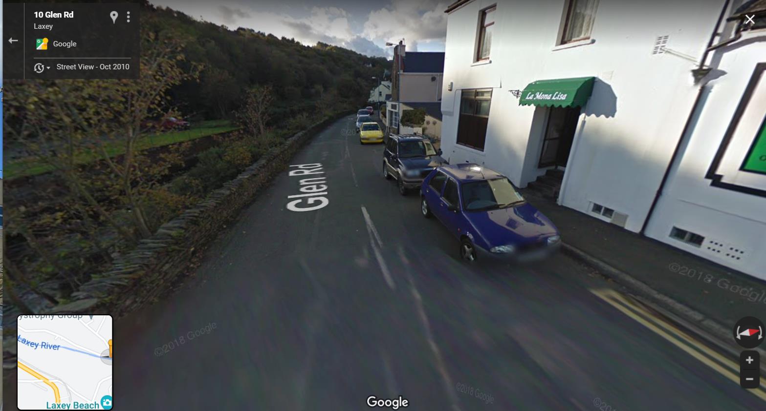 A Google Street View screenshot showing a road scene on Glen Rd in Laxey, featuring parked cars, a white building with a green awning, and a stone wall along the roadside.