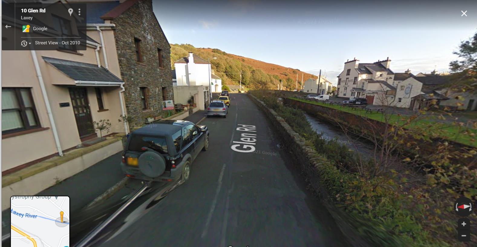 Google Street View screenshot showing a road scene in Laxey with a stone wall along the river and buildings on the left.