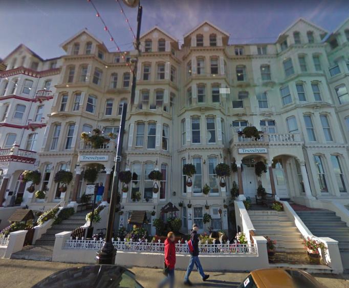 A street-level photograph showing a row of large, multi-story terraced buildings with pastel facades and hanging flower baskets, likely a hotel or guest house.