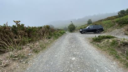 A photograph showing a gravel access track leading up a misty, rural hillside with a black SUV parked on the right.