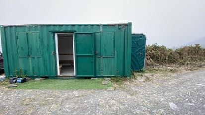 A photograph showing a green shipping container structure situated on a gravel and grass verge, likely representing the proposed storage or clubhouse facility.