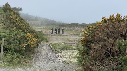 A photograph showing a rural outdoor site with a gravel track leading to a cleared area where people are standing near wooden posts.