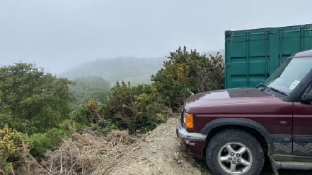 A photograph showing a maroon SUV parked on a dirt track next to a large green shipping container in a misty rural setting.