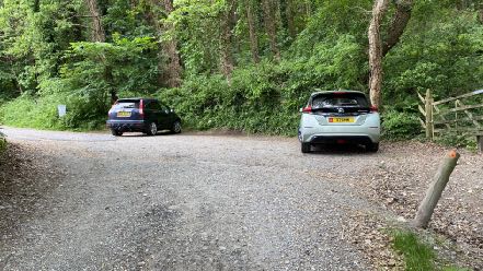 A photograph showing a gravel parking area with two cars parked in front of a dense wooded area.