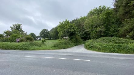 A photograph showing a rural road junction with a path leading into a grassy field surrounded by trees and hedgerows.