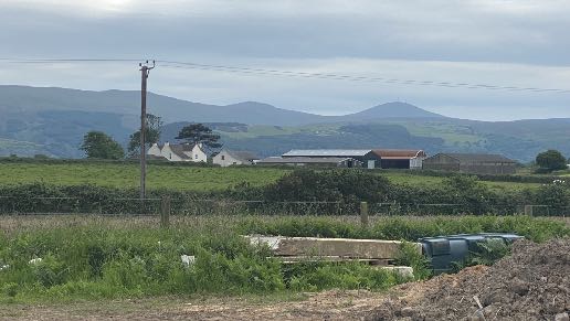 A landscape photograph showing a rural setting with rolling hills in the background and a row of white buildings and agricultural structures in the mid-ground.