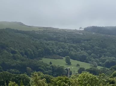 A photograph showing a rural, hilly landscape with dense woodland and green fields under an overcast sky.