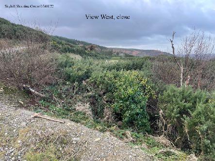 A photograph showing a grassy, vegetated slope with scrub and trees, labeled as a 'View West, close' from the Skyhill Shooting Centre.