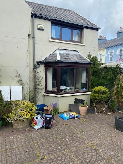 A photograph showing the rear exterior of a two-story house featuring a bay window and a paved patio area with gardening supplies.