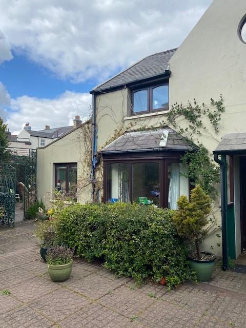 A photograph showing the exterior of a cream-colored residential house with a bay window, ivy, and a paved patio area with garden plants.