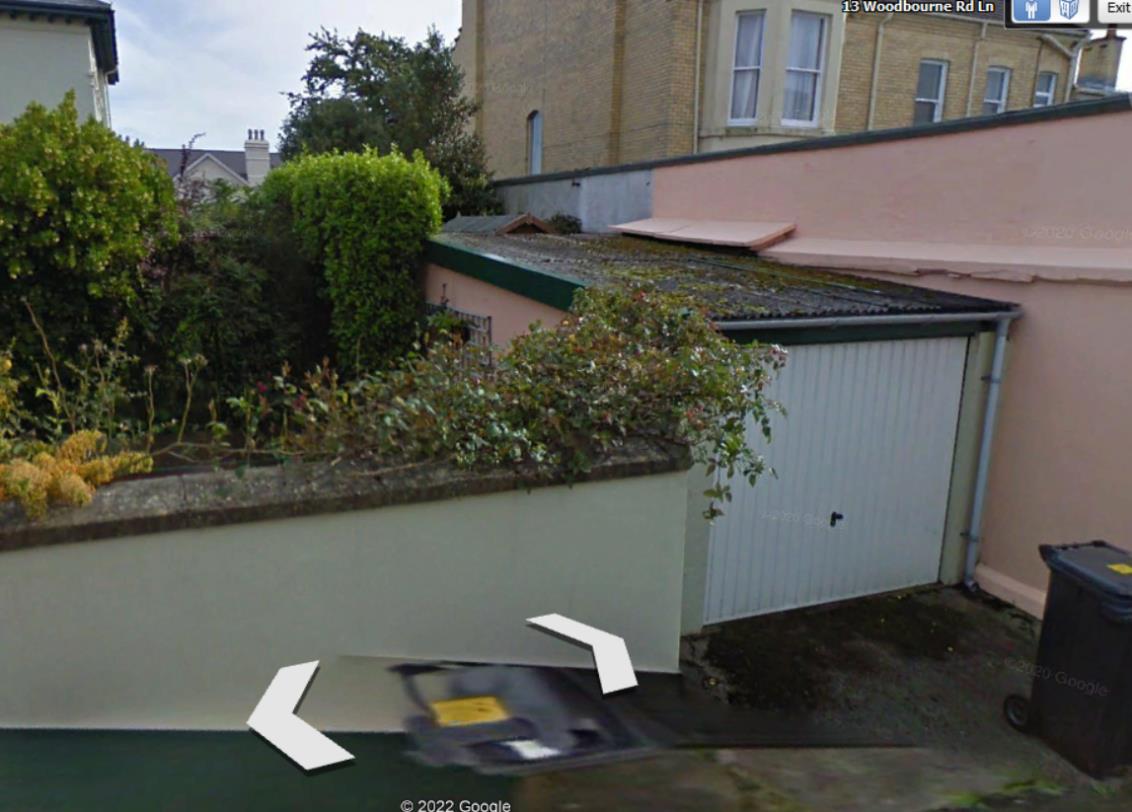A street-level photograph showing a residential property featuring a white garage door, a low boundary wall with vegetation, and a larger building in the background.