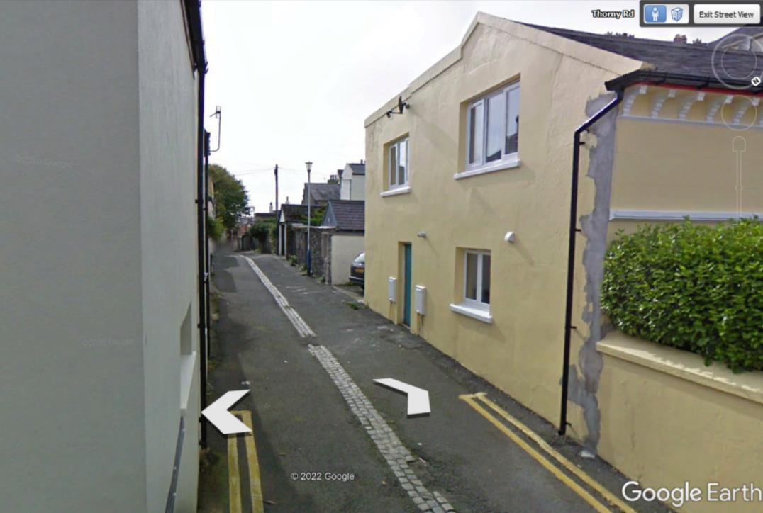 A Google Earth street-level screenshot showing a narrow residential road with a yellow two-story house on the right and a white wall on the left.