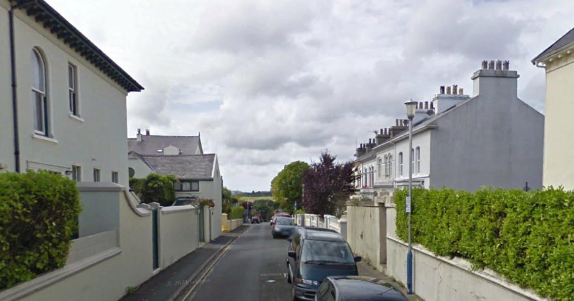 A street-level photograph showing a residential road in Douglas with terraced houses, parked cars, and boundary walls.